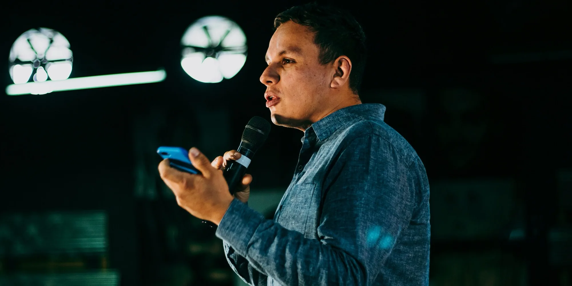A man in a denim shirt holding a mic and his phone, speaking out to a dark room
