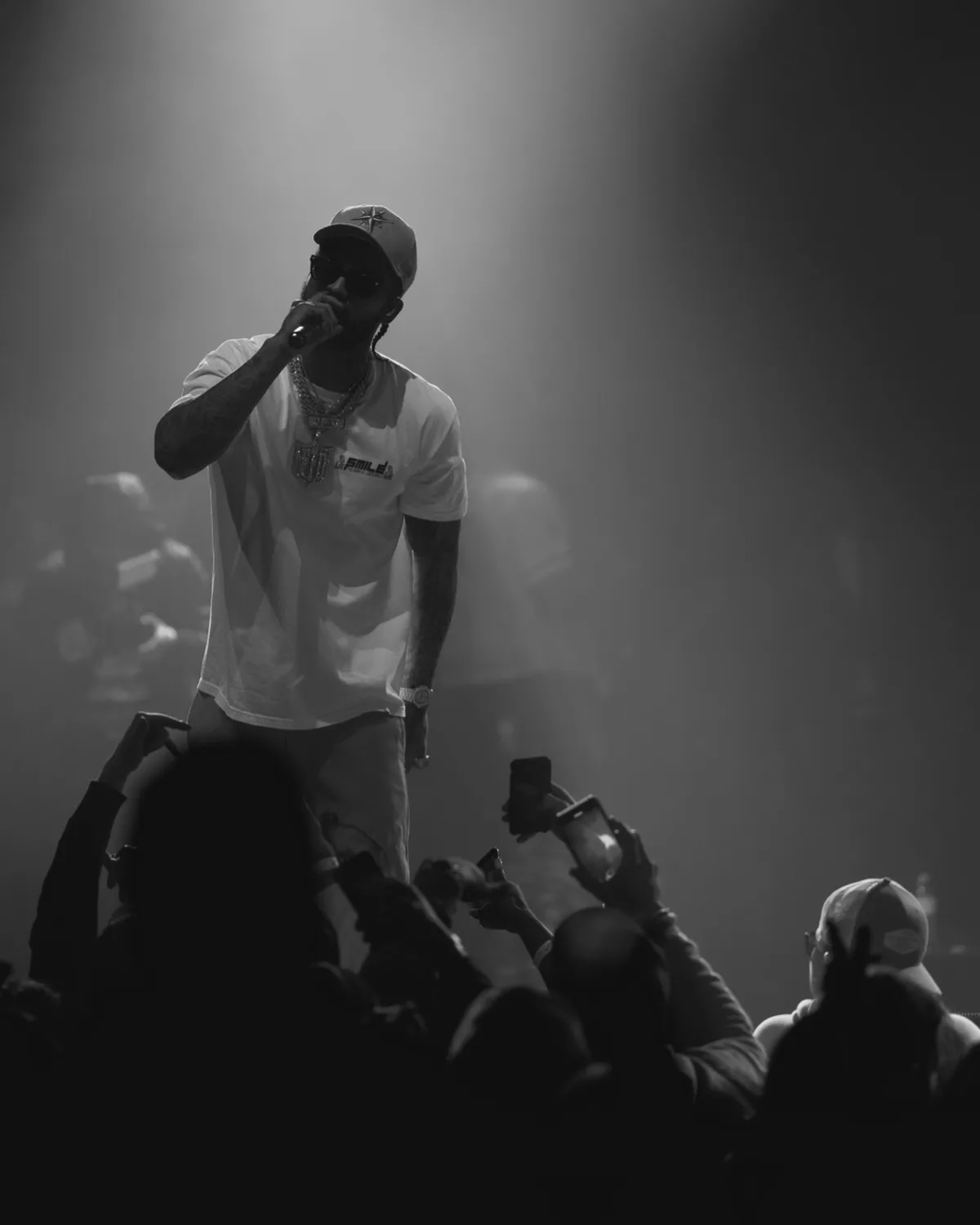 Black and white: a rapper under a spotlight, mic up, the crowd below holding phones in the air