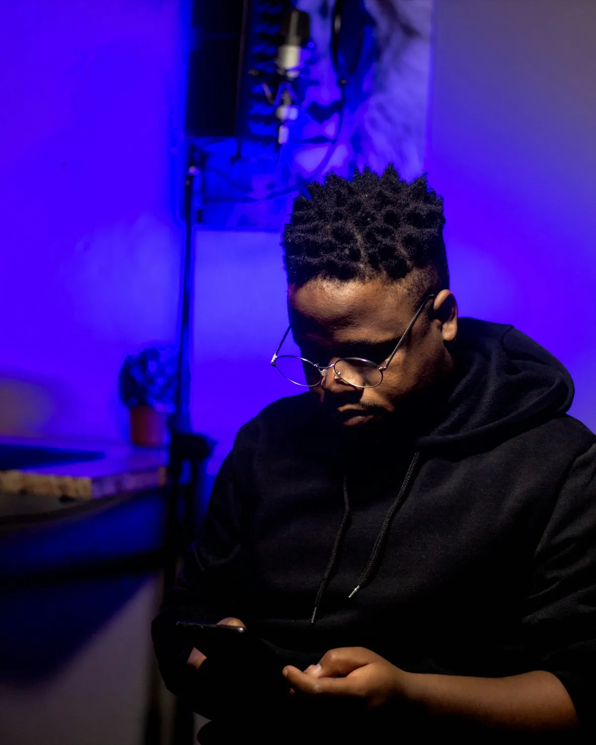 A writer in glasses reading his phone in a blue-lit room, a microphone on a stand behind him