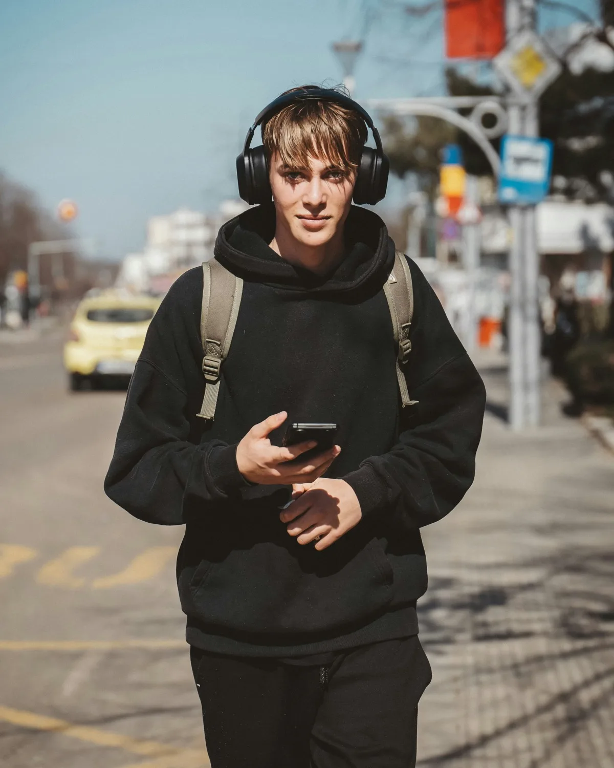 A young man in headphones and a black hoodie walking down a city sidewalk toward the camera, phone in hand