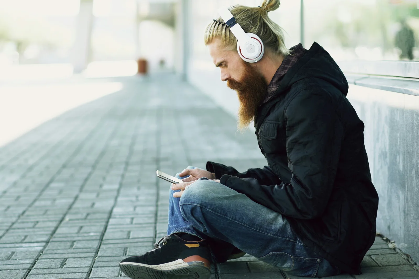 A writer sitting on a city sidewalk, hair tied back, headphones on, phone resting on his knee