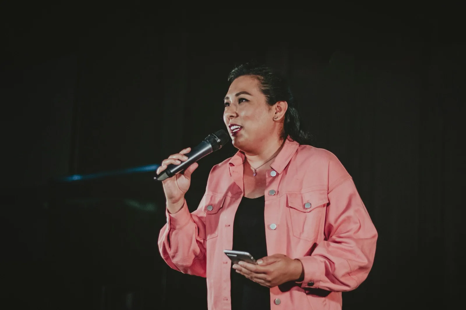 A woman in a pink jacket on a dark stage, mic in one hand, phone in the other, talking to the room