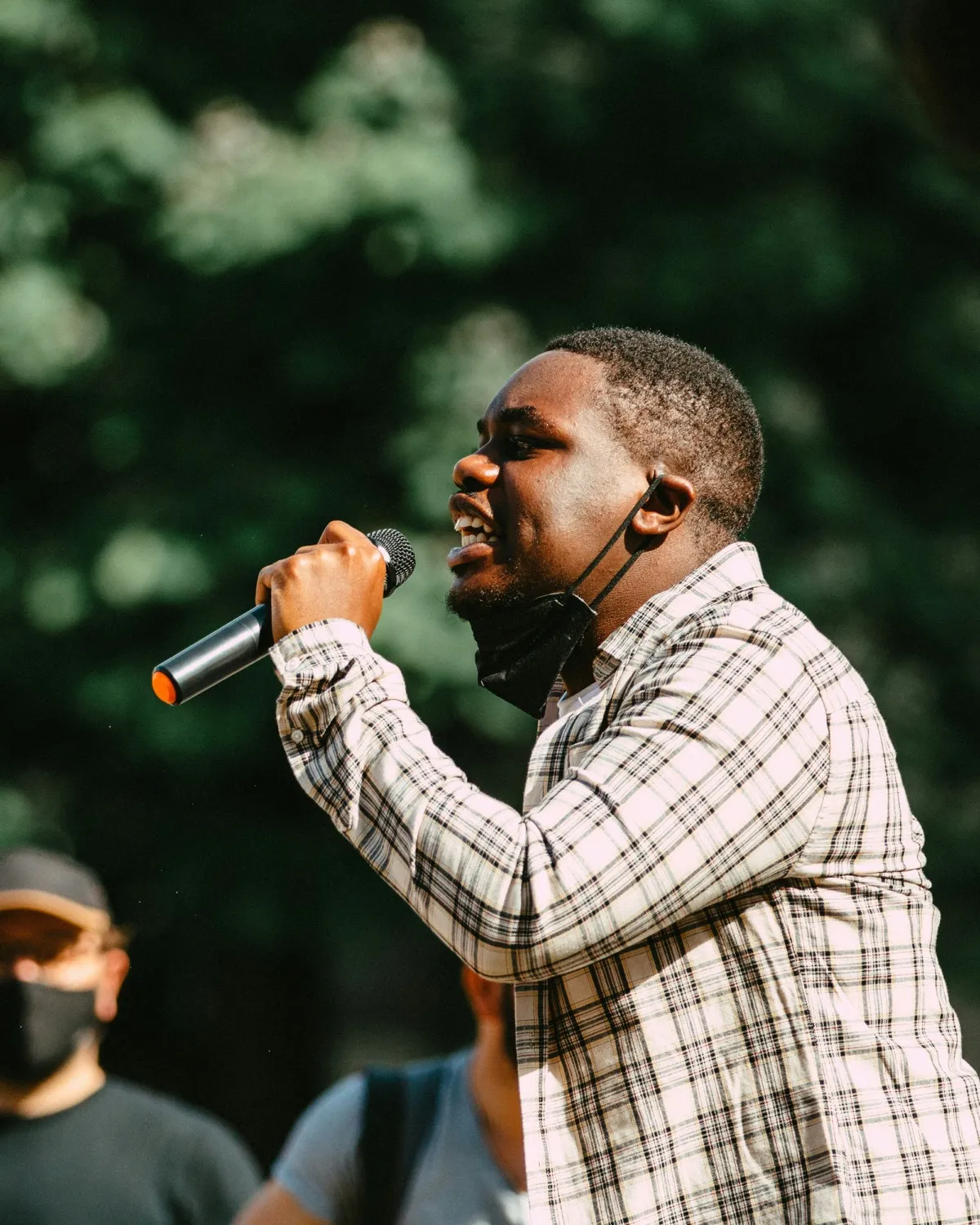 A man in a plaid shirt rapping into a mic outdoors, a crowd behind him