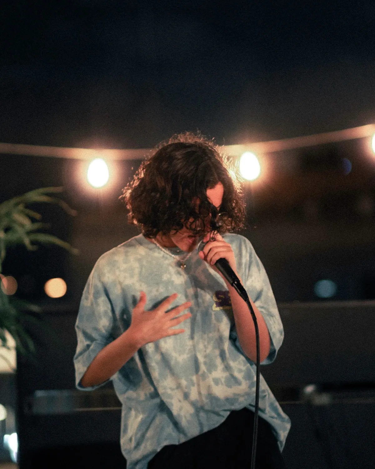 A performer on a rooftop at night, curls over the face, mic to the mouth, string lights behind